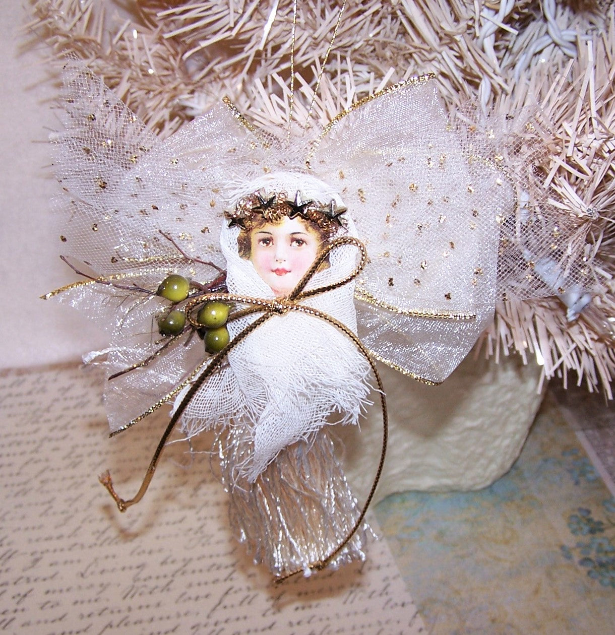 An angel ornament made of tulle, gauze, ribbon, cord and a tassel in colors of gold & cream. Backed with a wood heart which can be personalized by the new owner