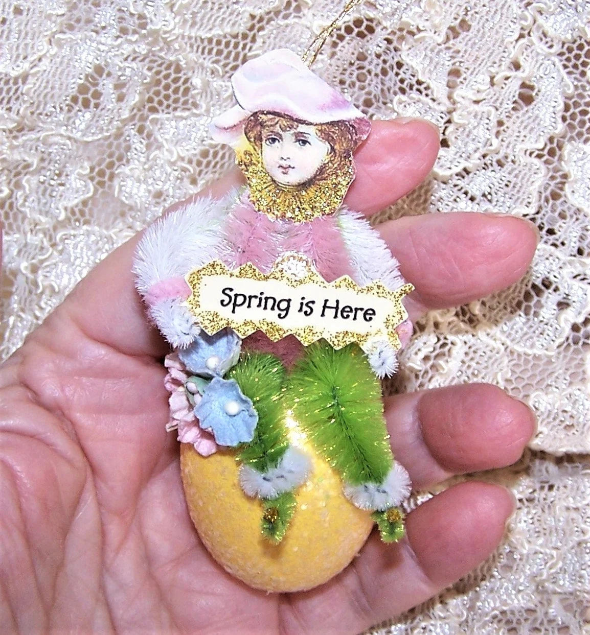 Handmade chenille ornament designed in a Victorian Revival manner. A little girl sits on a sparkly Easter Egg and holds a sign that reads SPRING IS HERE.