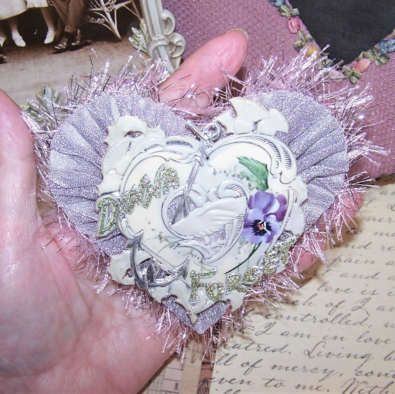 Heart shaped Antique Victorian celluloid token over a dusty lavender fabric heart. Sentiment is DINNA FORGET aka Forget Me Not. Handpainted pansy graphics.