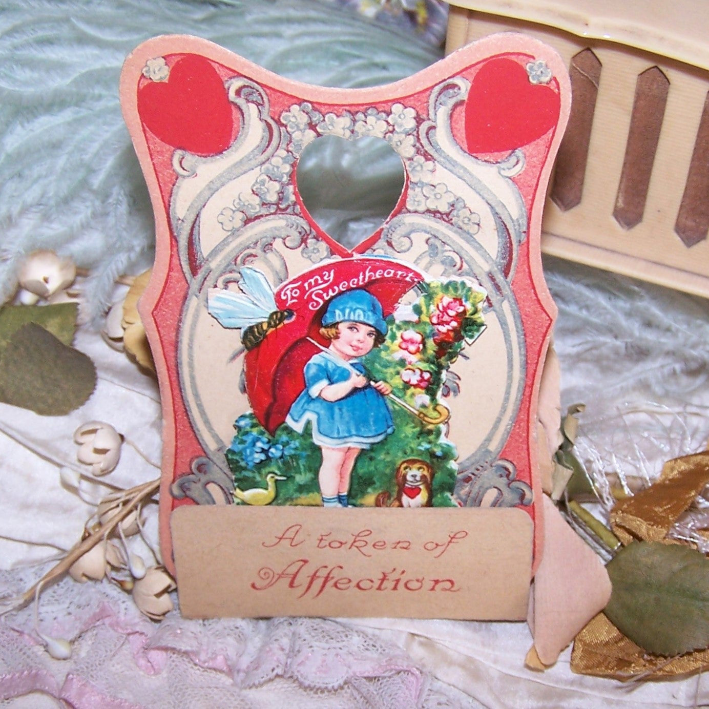 Used Valentines Day fold down card - printed in Germany.  Sentiment is A TOKEN OF AFFECTION.  Chromo die cut of girl carrying an umbrella in front of flowers.  