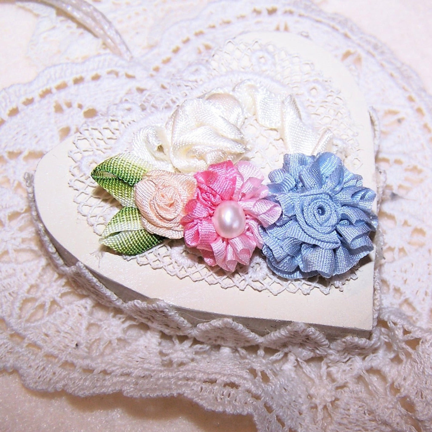 Entirely hand sewn, here's a French Revival ribbon applique composed of a trio of ribbon roses.  The hand rolled florals are made of 100% silk ribbon in colors of baby pin, baby blue and cream.  A genuine freshwater cultured pearl sits at the center of the pink flower.  Ombre green ribbon leaves complete the piece.  Length is 1-5/8"; width at the blue floral is 3/4".  