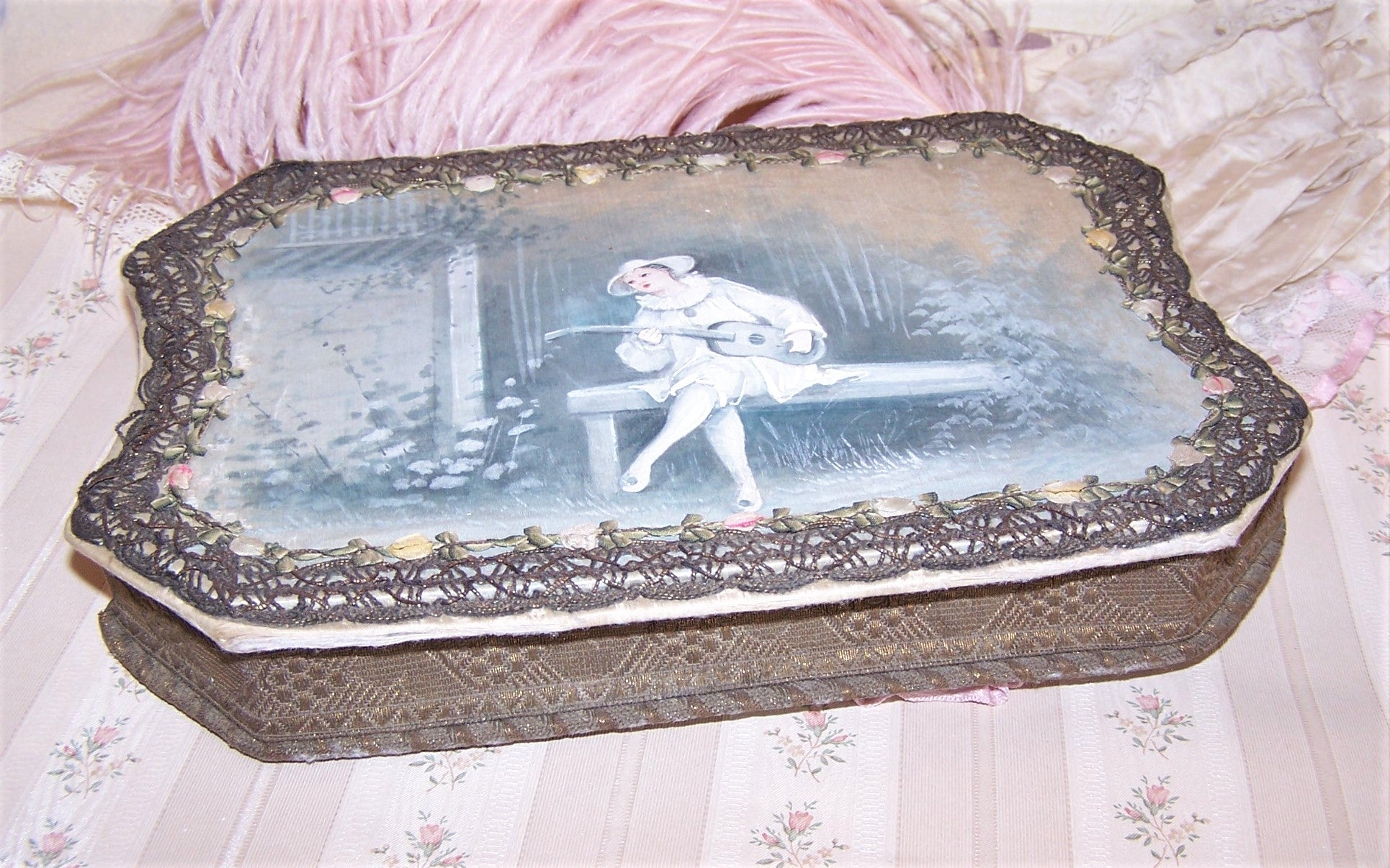 Antique French candy box handpainted with the image of Pierrot sitting in a garden.  The box is covered in braid and rococo ribbon roses trim. 
