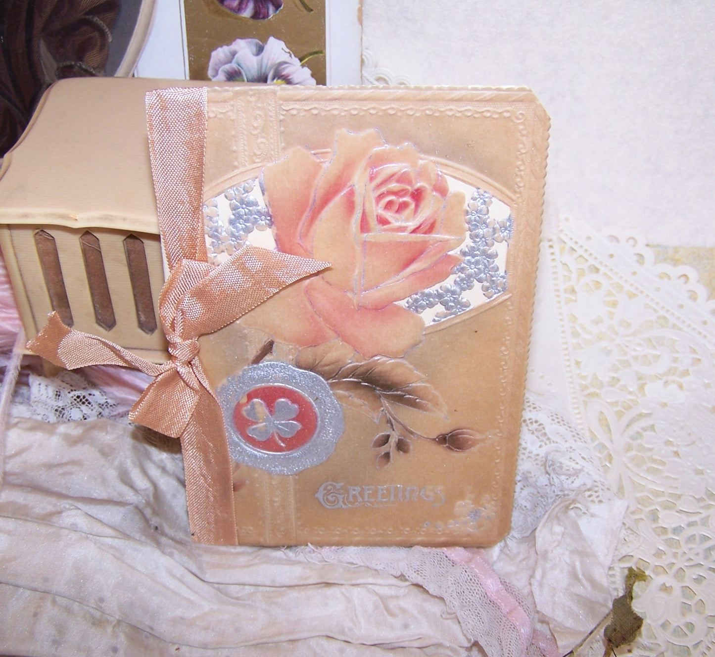 Christmas greeting card with an embossed celluloid cover of roses and 4 leaf clovers; the front sentiment is GREETINGS. Open the card to reveal a clean interior with a small Christmas poem. 