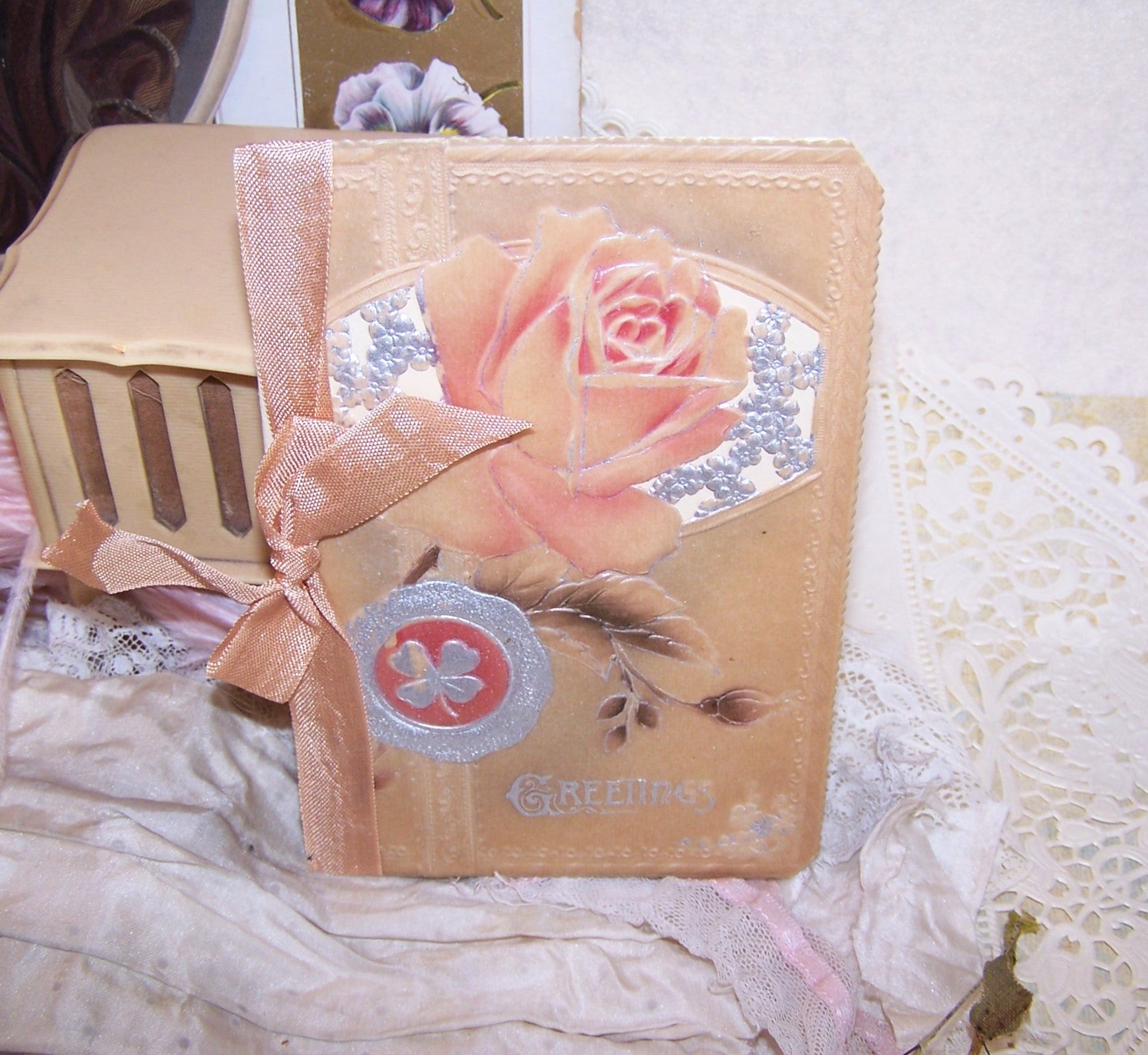 Christmas greeting card with an embossed celluloid cover of roses and 4 leaf clovers; the front sentiment is GREETINGS. Open the card to reveal a clean interior with a small Christmas poem. 