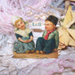 Antique Victorian die cut printed by L&B, the design number is 3847 and the image is of a Dutch Boy & Girl sitting on the ground. 2-1/2" wide x 2-11/16".