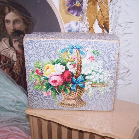 Antique paper box featuring a Victorian die cut of a basket of florals over a silver paper background.  Cotton batting on the inside.  3-11/16" x 3-3/16".