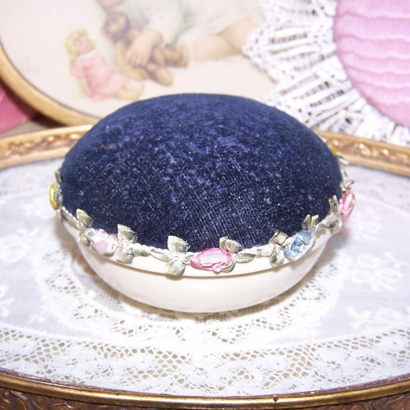 Used Art Deco pin cushion - celluloid base, blue velvet cushion and a length of French rococo ribbon at the center. Height is about 1-3/4"; width is about 3". 