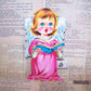 Cardboard hanging decoration of a small angel dressed in pink and white. Singing hymns or carols, the wall or tree piece can be used for Christmas or Easter. 