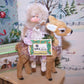 Original mixed media holiday ornament. My pink chenille girl sits on a brown fawn and holds a sign that reads GOOD WISHES.  5-1/2" x 3-1/2"