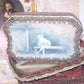 Antique French candy box handpainted with the image of Pierrot sitting in a garden.  The box is covered in braid and rococo ribbon roses trim. 
