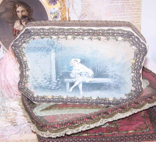 Antique French candy box handpainted with the image of Pierrot sitting in a garden.  The box is covered in braid and rococo ribbon roses trim. 