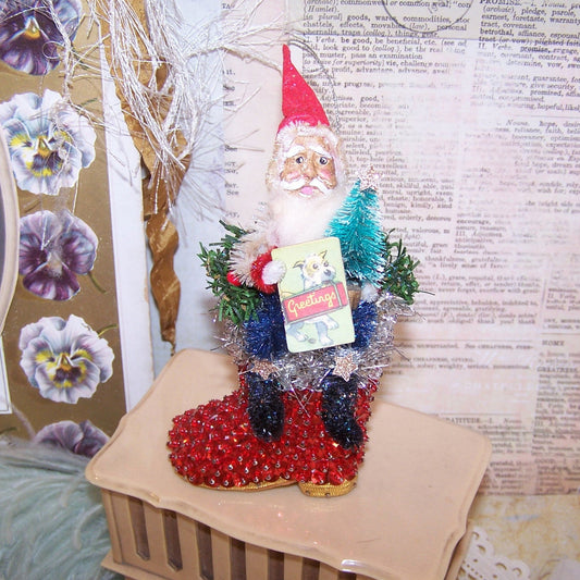 Handcrafted One of a Kind chenille & clay face Santa Claus sitting on a sequined boot ornament/mantel decoration. He holds a sign reading GREETINGS. 5.5" x 2.5"