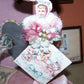 Handmade OOAK chenille ornament on vintage greeting card.  A baby girl sits on the top edge of a card meant to accompany a baby shower gift.  Pink coloring. 