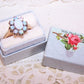 Light blue paper ring box with slotted insert on the inside and Antique Victorian die cut reading WITH MY LOVE to the top. 1-1/4" x 15/16" x 1-3/16" high. 